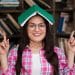 Mulher jovem usando óculos e camisa xadrez rosa com livro verde aberto sobre a cabeça em biblioteca.