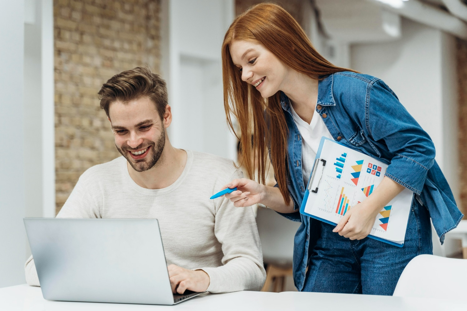 Homem e mulher jovens trabalhando juntos em gráfico no laptop em ambiente de escritório moderno.