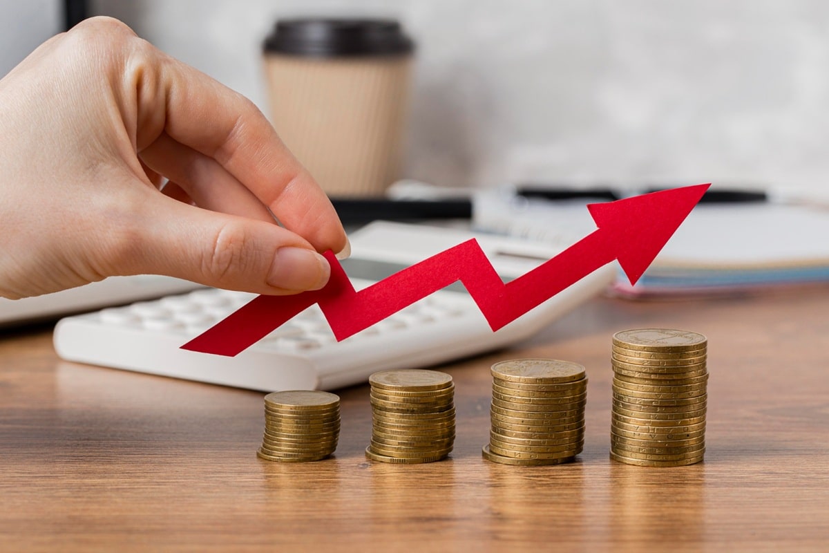 Mão segurando seta vermelha ascendente sobre pilhas crescentes de moedas douradas representando crescimento de investimentos
