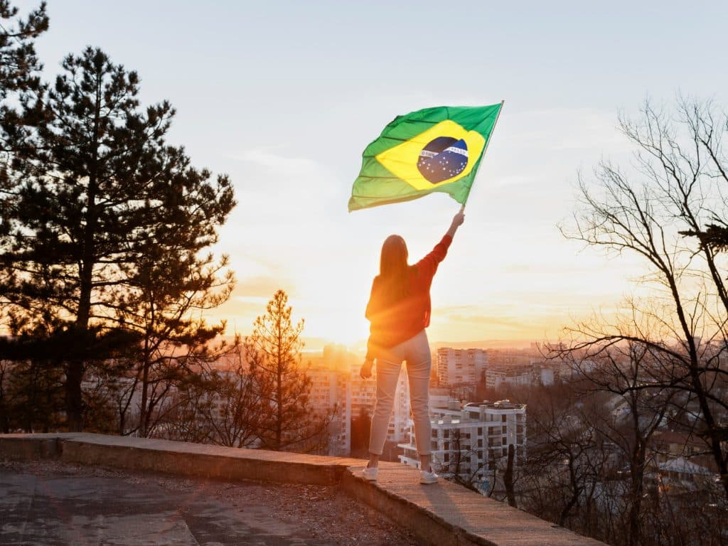 Mulher de cabelo longo segurando bandeira do Brasil em um muro com cidade ao fundo e pôr do sol.