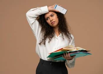 Mulher preocupada segura vários livros e olha para celular mostrando gráfico na tela.