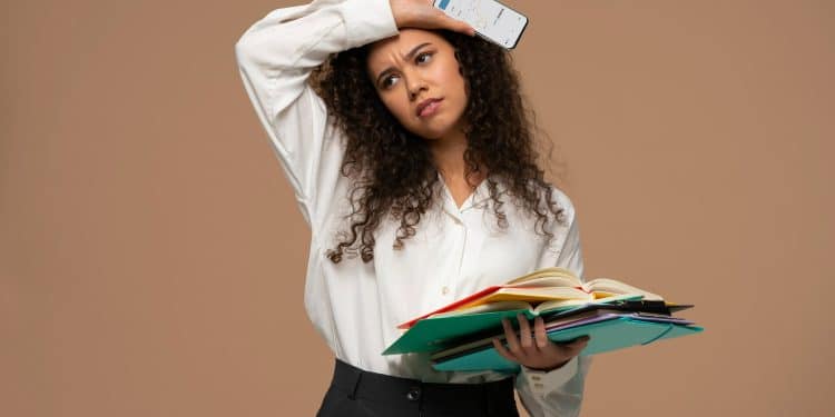 Mulher preocupada segura vários livros e olha para celular mostrando gráfico na tela.
