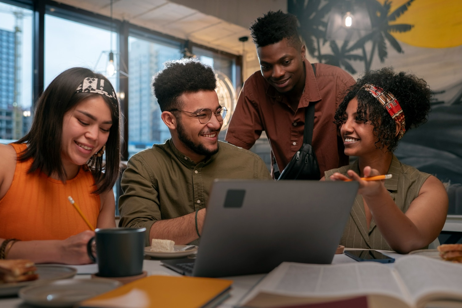 Grupo de quatro jovens sorrindo e colaborando, estudando juntos com notebook em ambiente moderno.