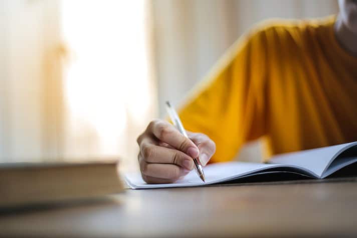 Imagem de uma mão de estudante segurando uma caneta e escrevendo em um caderno de anotações, com um livro ao lado.