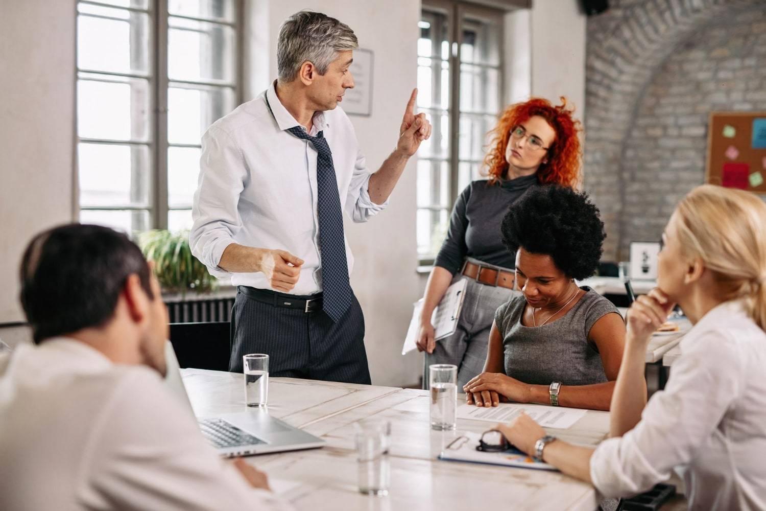Homem de terno falando gesticulando em reunião de negócios com equipe atenta em escritório moderno.