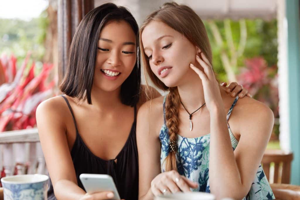 Duas jovens sentadas juntas olhando para um smartphone em ambiente ao ar livre com plantas ao fundo.