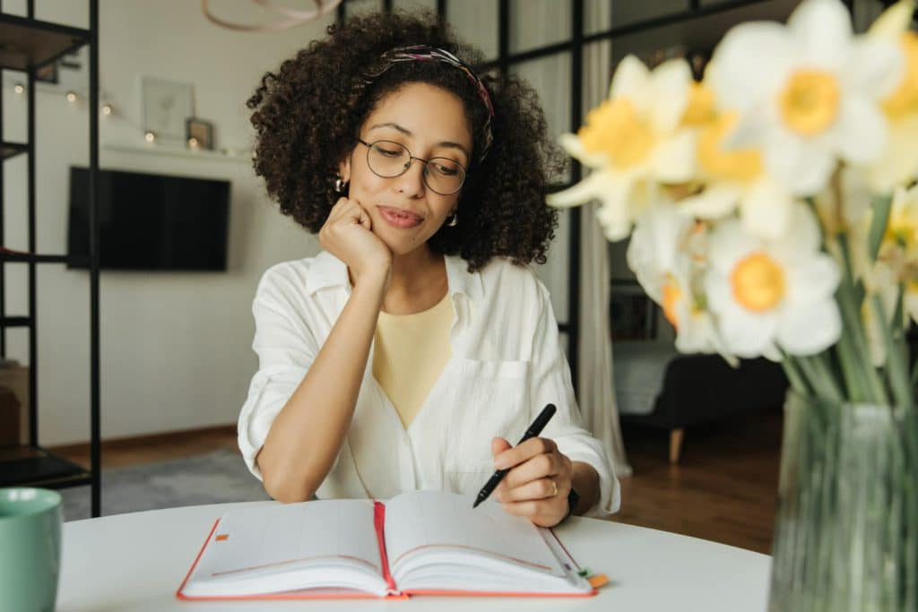 Mulher usando óculos, com camisa branca, escrevendo em caderno aberto sentada à mesa com flores ao lado