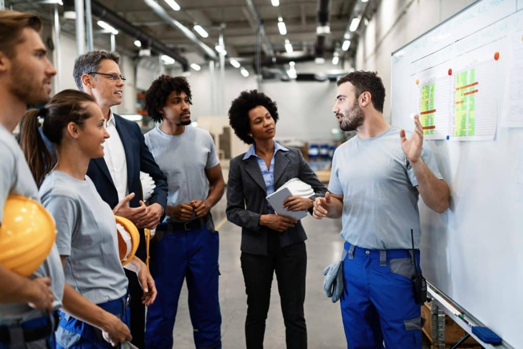 Grupo de trabalhadores e gestores em indústria, discutindo planejamento diante de quadro branco com gráficos.