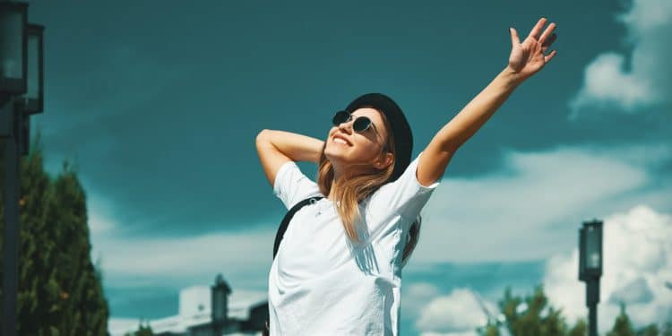 Mulher sorridente com óculos escuros e chapéu preto celebrando ao ar livre em um dia ensolarado.