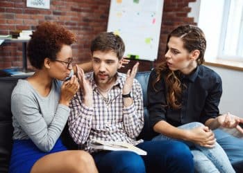 Três jovens sentados em sofá discutindo de forma intensa, com uma mulher falando, um homem gesticulando e outra mulher surpresa.
