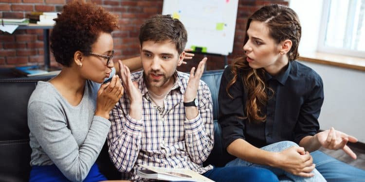 Três jovens sentados em sofá discutindo de forma intensa, com uma mulher falando, um homem gesticulando e outra mulher surpresa.