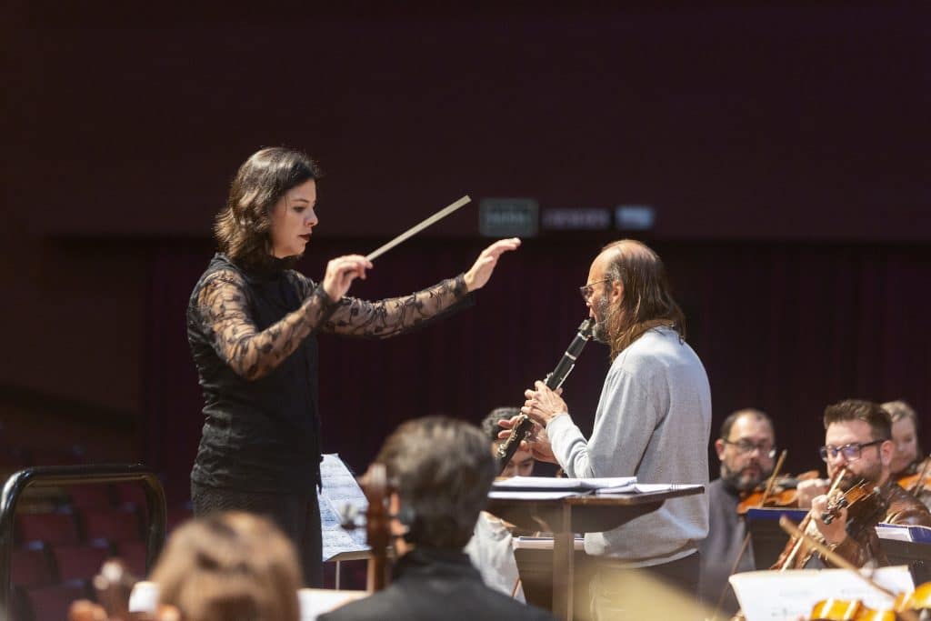 Mulher regente de orquestra com batuta e um músico tocando clarinete durante uma apresentação ou ensaio.