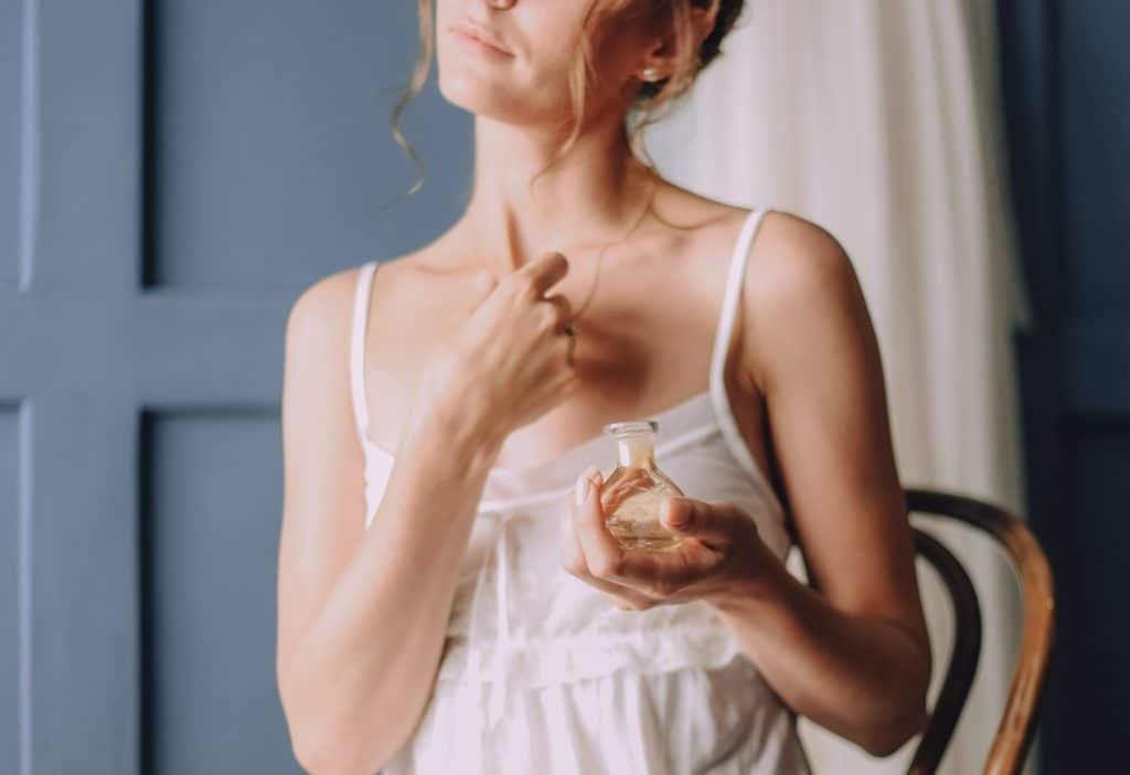 Mulher em vestido branco segurando um pequeno frasco de perfume em ambiente interno.