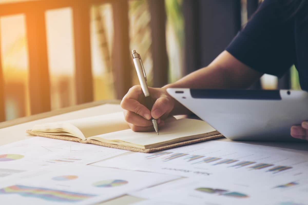 Pessoa fazendo anotações em caderno com gráficos e relatórios estatísticos sobre mesa de estudo iluminada
