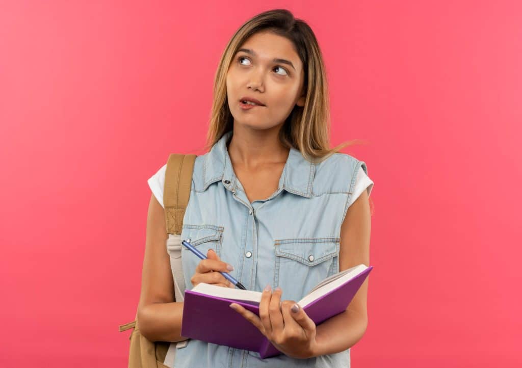 Jovem estudante segurando caderno roxo e caneta, olhando para cima, vestida com colete jeans frente rosa.