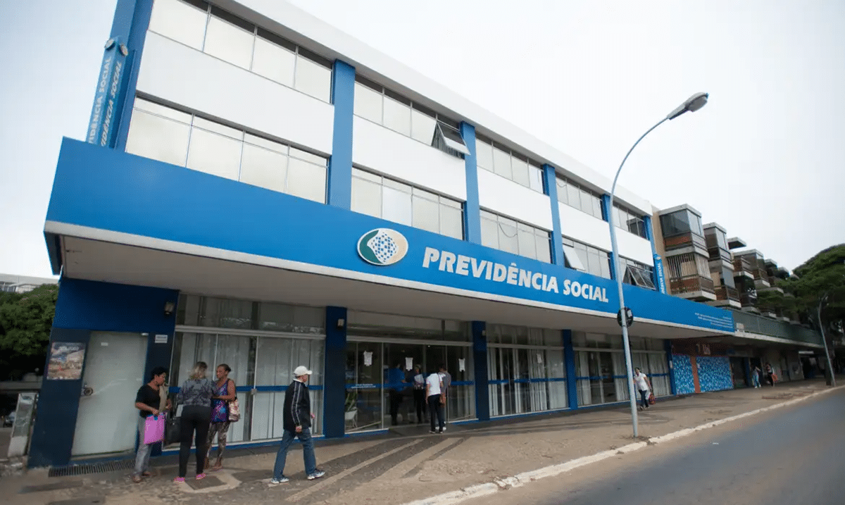 Fachada de agência da Previdência Social azul e branca com beneficiários aguardando atendimento presencial em calçada
