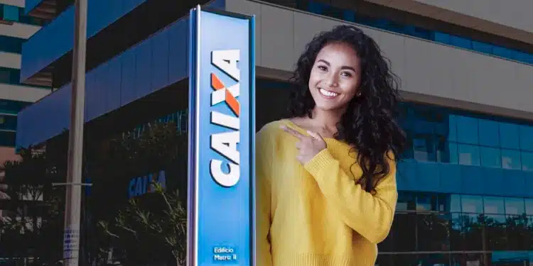 Mulher sorridente com blusa amarela e calça jeans, apontando para a placa da Caixa Econômica Federal, simbolizando o anúncio do novo concurso público e o andamento do processo seletivo