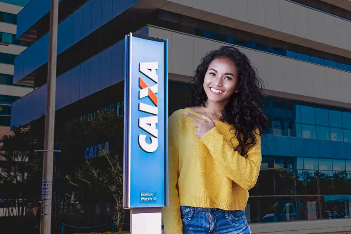 Mulher sorridente com blusa amarela e calça jeans, apontando para a placa da Caixa Econômica Federal, simbolizando o anúncio do novo concurso público e o andamento do processo seletivo