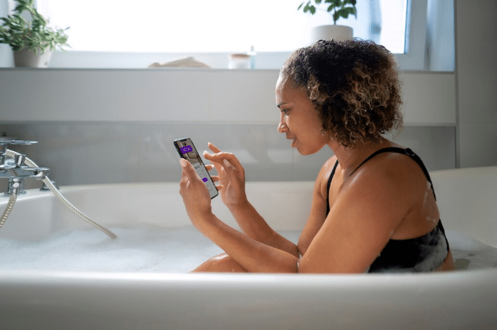 Mulher de cabelos cacheados em uma banheira cheia de espuma, usando o celular.