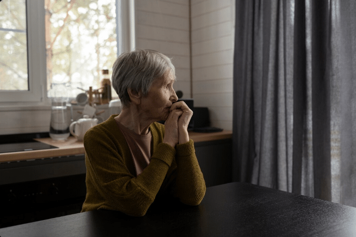  Mulher idosa, sentada sozinha à mesa de uma cozinha, com uma expressão de preocupação e mãos juntas, representando os desafios emocionais da doença de Alzheimer.