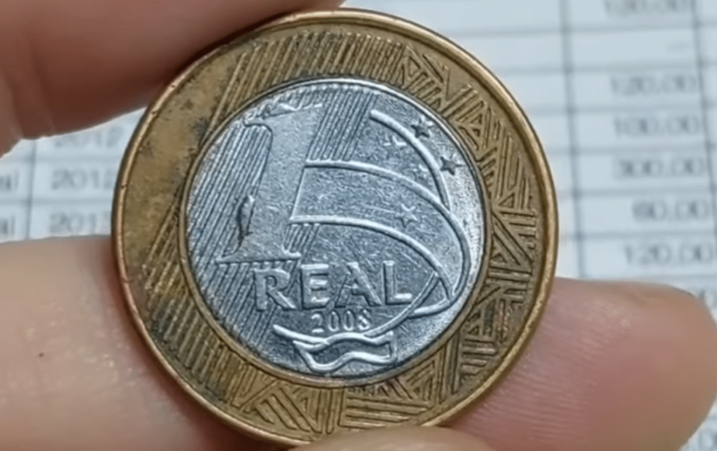 Moeda de um real do ano de 2008, mostrando os detalhes do valor facial e a constelação do Cruzeiro do Sul, sendo segurada entre dois dedos com um fundo desfocado de uma tabela de preços numismáticos.
