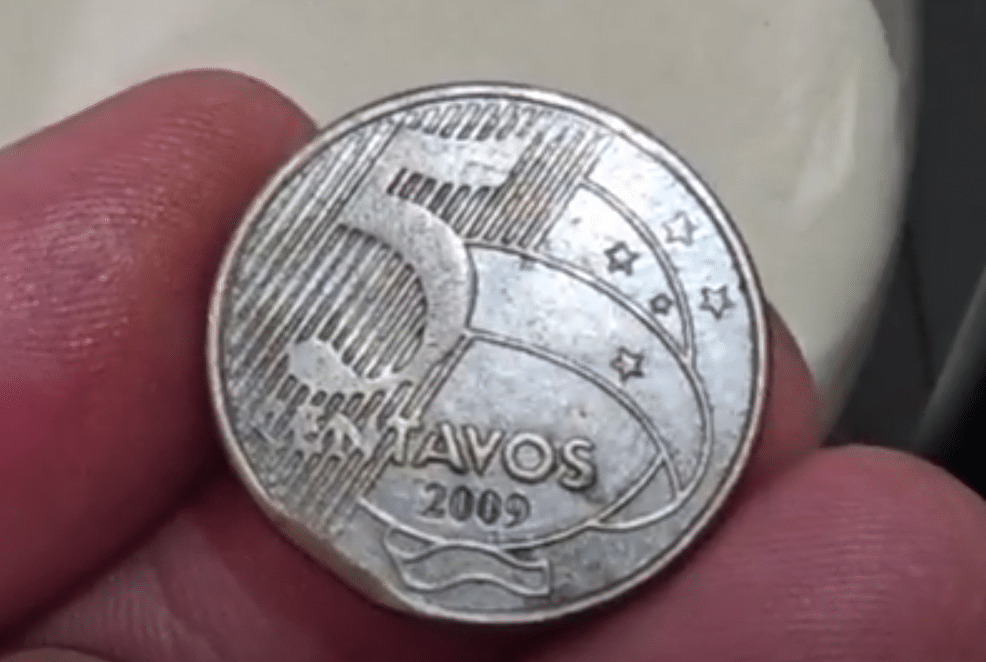 Uma mão segurando uma moeda de 5 centavos do ano de 2009 com um defeito na borda, o que a torna valiosa para colecionadores. A imagem serve para ilustrar um dos erros de cunhagem.