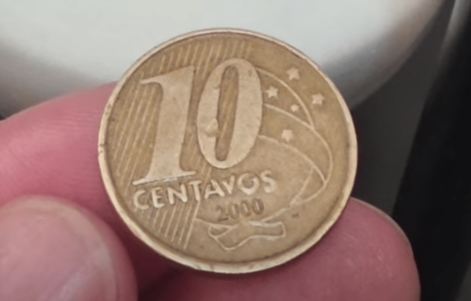 Uma moeda de 10 centavos do ano 2000, segurada entre os dedos de uma pessoa, destacando a cunhagem com um possível defeito de fabricação.