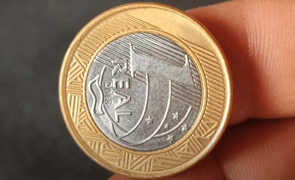 Moeda de 1 real de 2018 em close-up, mostrando o erro de cunhagem onde o reverso (o rosto da República) está de lado, simbolizando uma anomalia rara.