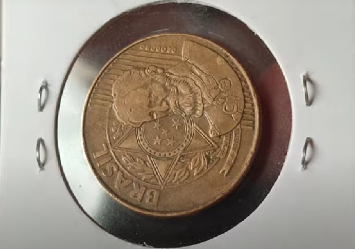 Detalhe ampliado do reverso de uma moeda de 25 centavos (provavelmente de 2008) guardada em um suporte, destacando o erro de cunhagem conhecido como reverso horizontal, que deve ser identificado girando a peça.