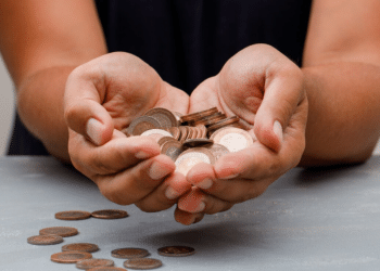 Mãos segurando um punhado de moedas e notas de real, com outras moedas espalhadas sobre uma mesa. A imagem representa o valor escondido nas moedas do dia a dia.
