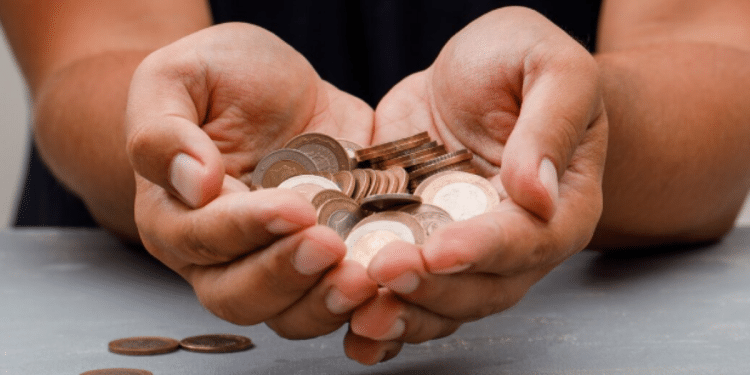 Mãos segurando um punhado de moedas e notas de real, com outras moedas espalhadas sobre uma mesa. A imagem representa o valor escondido nas moedas do dia a dia.