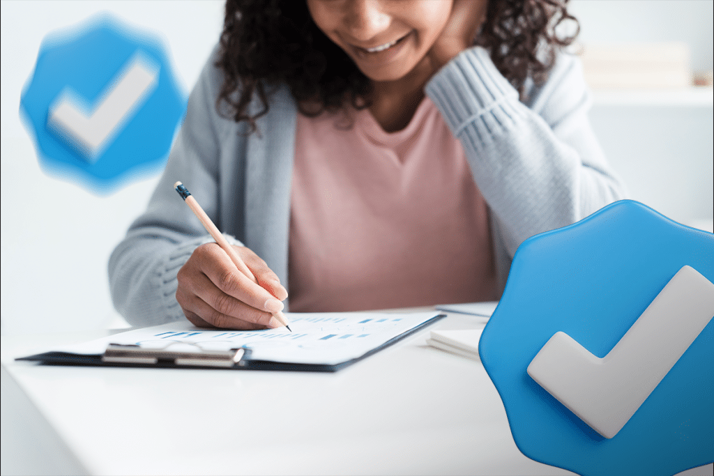  Mulher sorrindo, sentada à mesa e escrevendo em um documento, com ícones de verificação (checkmarks) em azul flutuando, representando a organização e a satisfação de quem se prepara para um concurso público.