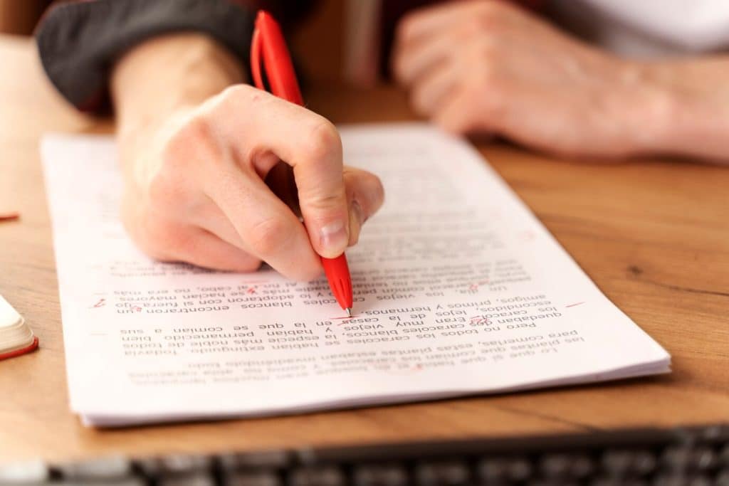 Mão segurando caneta vermelha revisando texto em documentos com anotações e correções.