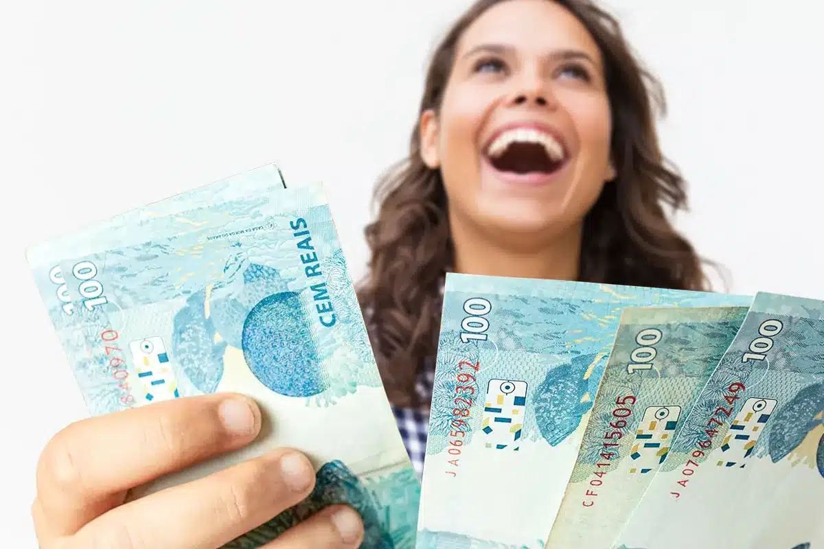 Jovem mulher sorridente, segurando e olhando para notas de R$ 100 reais, simbolizando a alegria e a oportunidade de receber o saque de até R$ 6.220 da CAIXA.