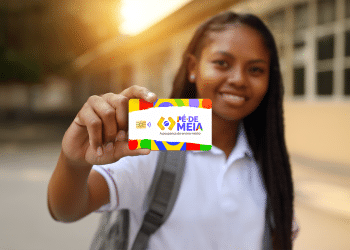 Estudante sorridente segurando cartão colorido do programa Pé-de-Meia com escola ao fundo em luz dourada