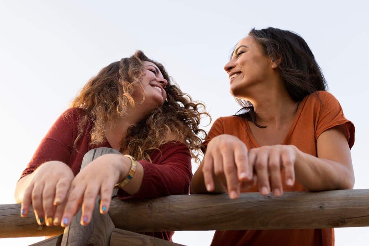 Duas mulheres sorridentes conversando apoiadas em corrimão de madeira ao ar livre ao pôr do sol