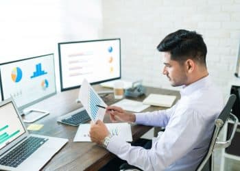 Analista de dados trabalhando em seu escritório, examinando relatórios de vendas em um ambiente de home office com dois monitores.