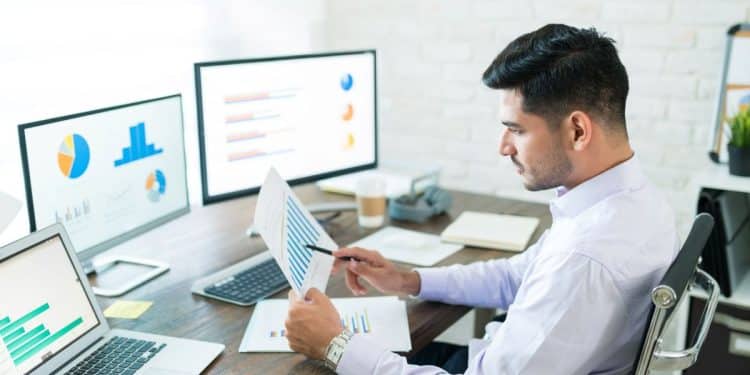 Analista de dados trabalhando em seu escritório, examinando relatórios de vendas em um ambiente de home office com dois monitores.