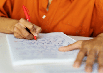 Mão segurando caneta vermelha e escrevendo em caderno pautado com escrita manuscrita azul e vermelha