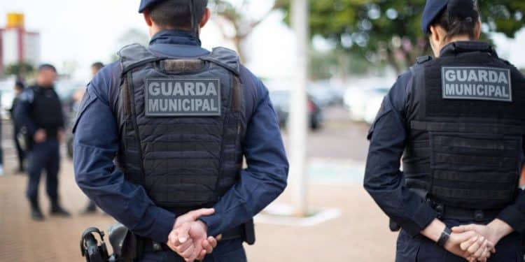 Dois guardas municipais com coletes pretos e uniformes azuis estão de costas em uma área externa urbana