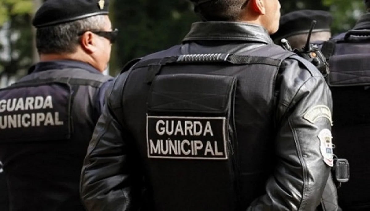 Três guardas municipais com coletes pretos e uniformes em movimento durante operação.