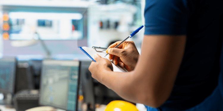 Profissional com uniforme azul escrevendo em prancheta em ambiente industrial com computadores e capacete amarelo.