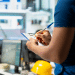 Profissional com uniforme azul escrevendo em prancheta em ambiente industrial com computadores e capacete amarelo.
