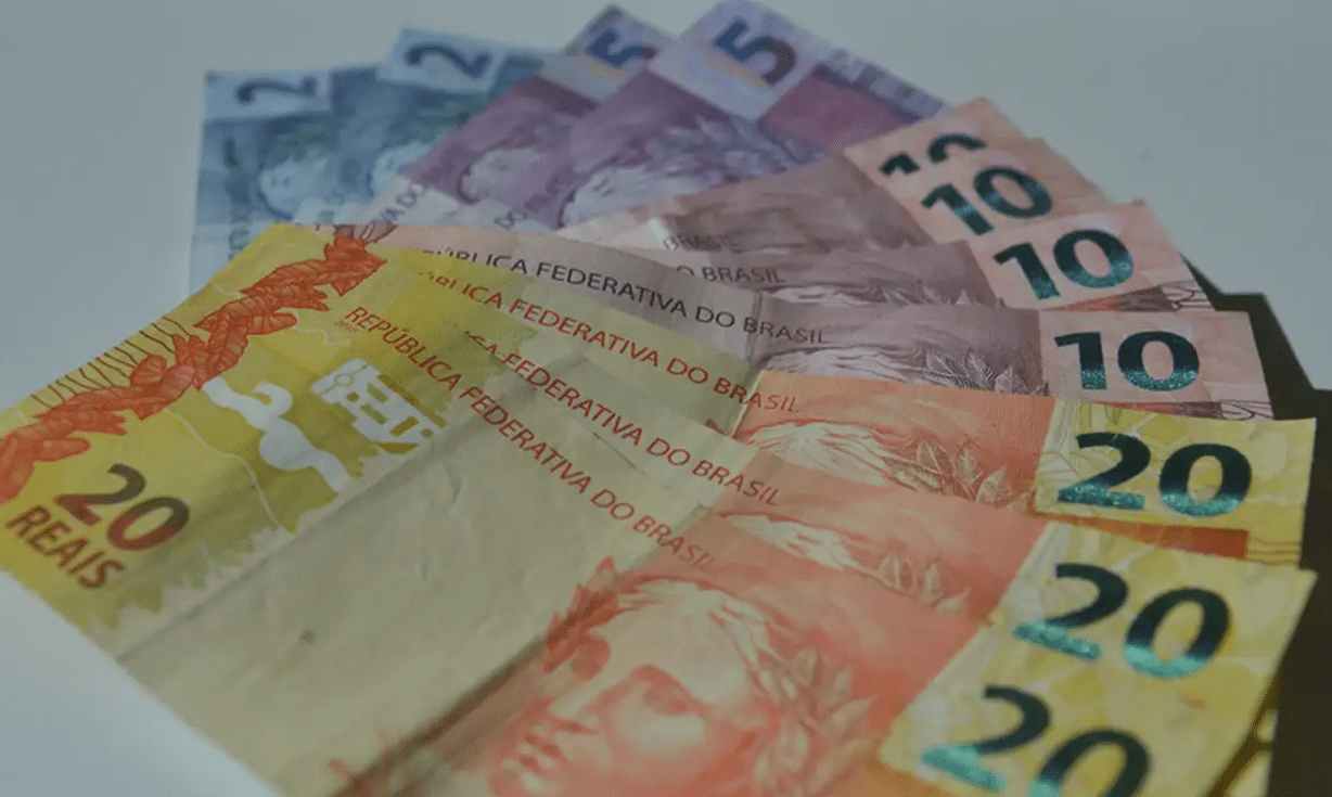 Notas de reais brasileiros em cédulas de 5, 10 e 20 espalhadas sobre superfície branca.