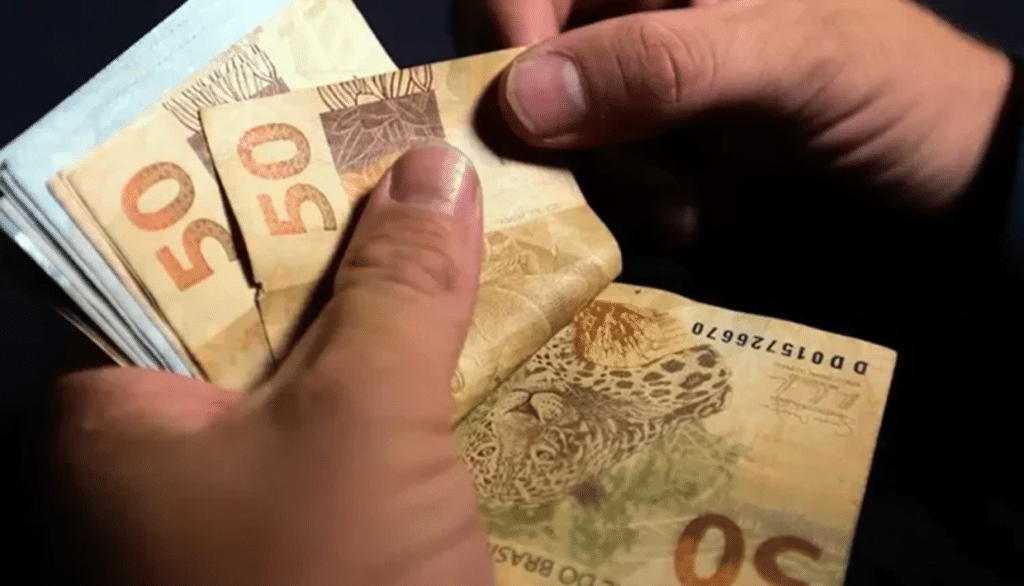 Mãos masculinas contando notas de cinquenta reais brasileiras com fundo escuro.
