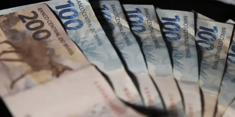 Cédulas de real brasileiro em valores de 100 e 200 distribuídas sobre fundo escuro.