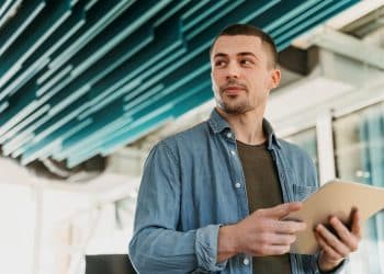 Jovem empreendedor com camisa jeans, segurando um tablet em um escritório moderno, simbolizando o planejamento de um novo negócio de franquia no interior.