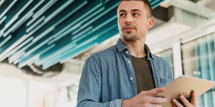 Jovem empreendedor com camisa jeans, segurando um tablet em um escritório moderno, simbolizando o planejamento de um novo negócio de franquia no interior.