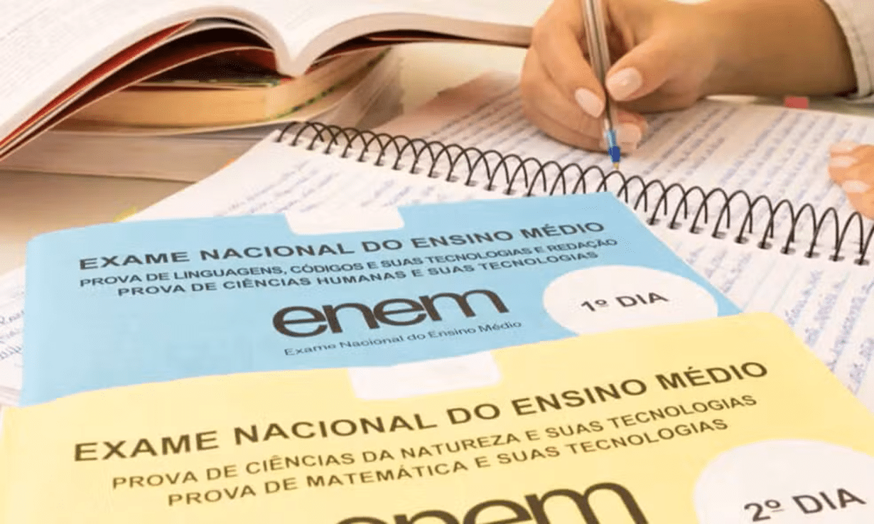 Cadernos do ENEM 1º e 2º dia sobre mesa com pessoa escrevendo em caderno espiral.