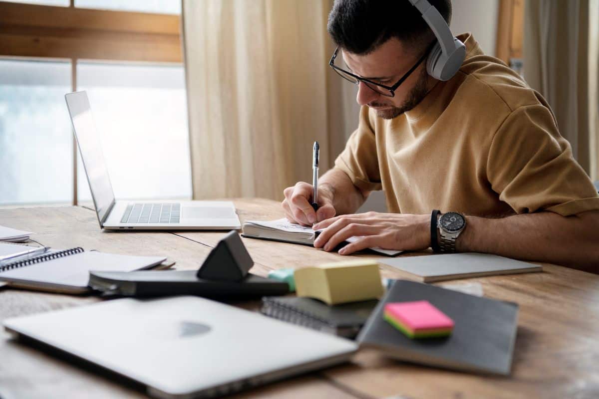 Homem com fones de ouvido estudando para concursos e fazendo anotações em um caderno, com um notebook ao lado.